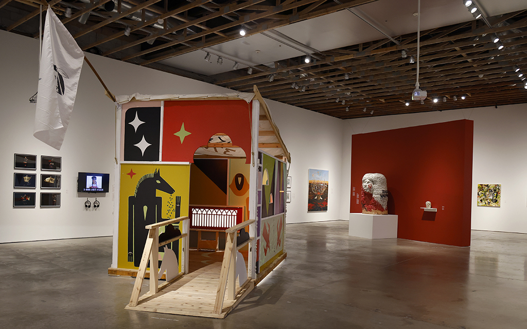 Interior of a modern art gallery with diverse artworks, including a colorful house-like installation and multiple framed photographs. The installation is made of panels have geometric designs, including a black horse-like figure on a yellow background. The house has an archway entrance and a small wooden ramp in front. To the left of the installation, a white flag is hanging from a wooden beam.
