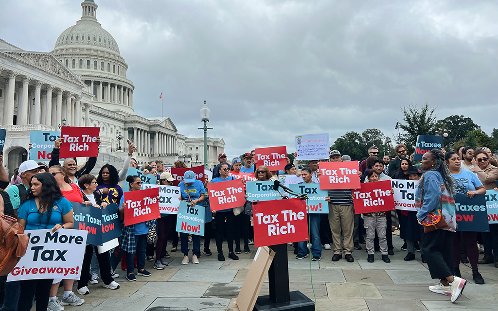 A crowd of people standing outside the capitol building holding signs that say "Tax The Rich."
