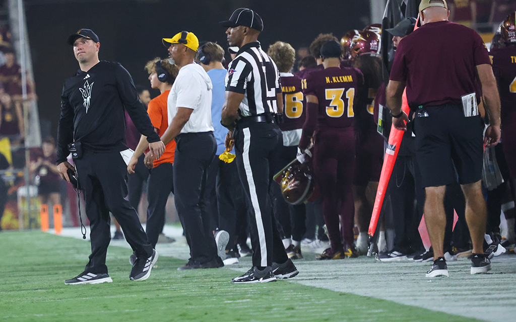 Football sideline with coaches, players in maroon uniforms, and an official in a striped uniform.