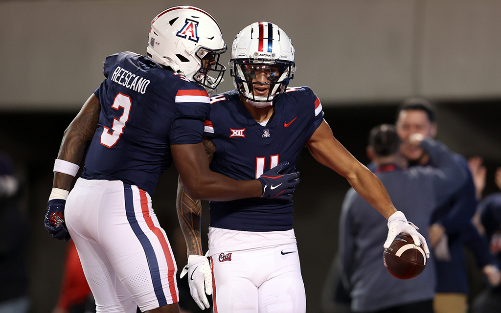 Two Arizona football players in blue uniforms celebrate on the field, one holding a football.
