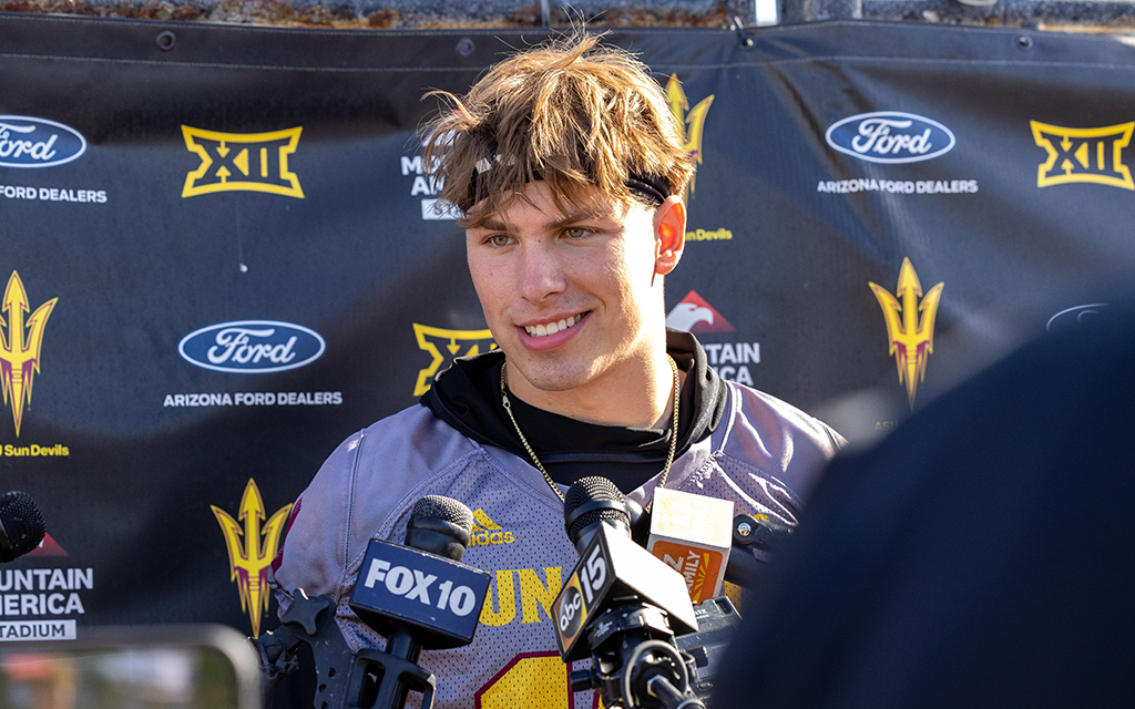Sam Leavitt in a sports jersey smiling during a press conference with microphones in front of him and a backdrop of sports logos.