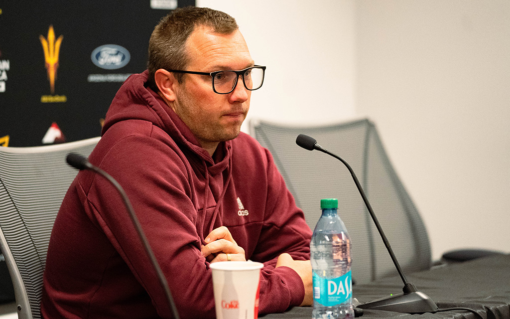Man in a maroon hoodie at a press conference, with a microphone, Coca-Cola cup, and Dasani water bottle on the table.