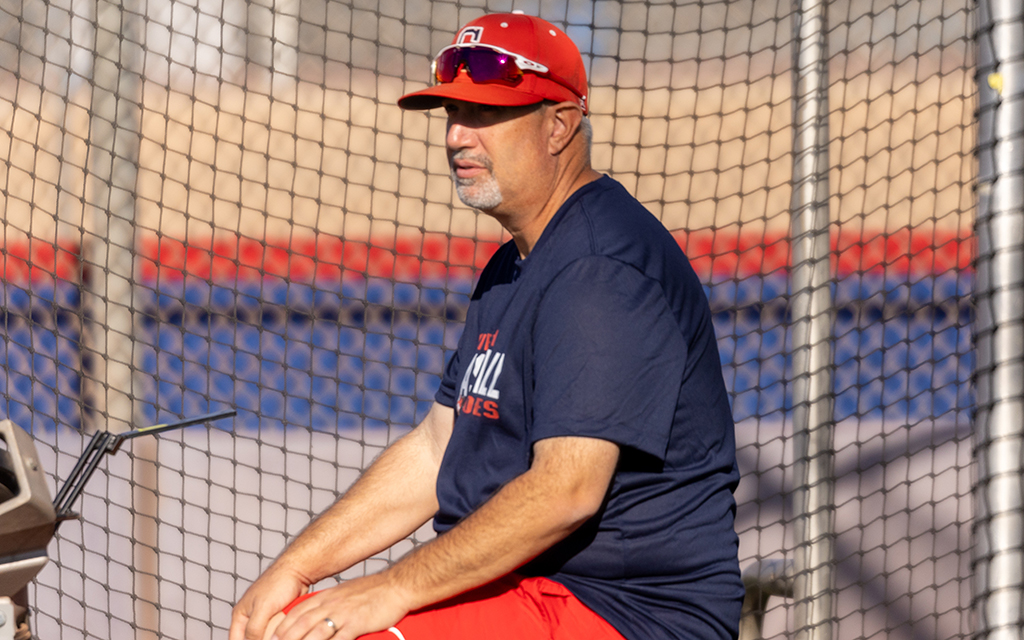 Jon Williams sitting by a baseball batting cage wearing a red cap and sunglasses.