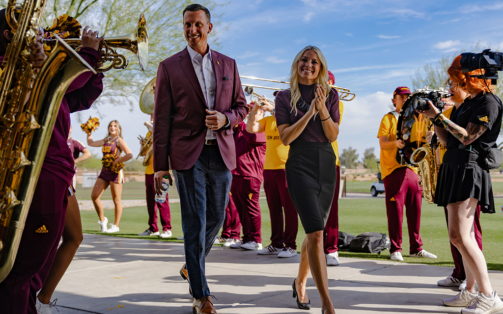 A man and woman walk amidst a marching band and cheerleaders in an outdoor setting.