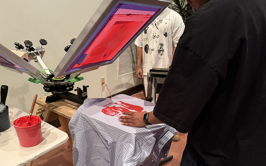 A person using a screen printing press to transfer a red design onto checkered fabric.