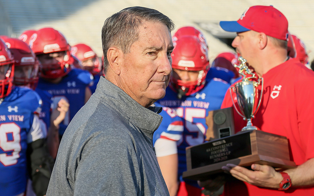 David Hines is in a gray jacket and stands in front of football players wearing blue uniforms and red helmets, with a trophy visible behind him.