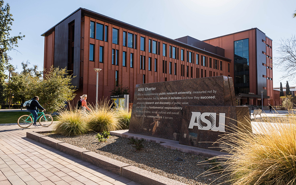 Arizona State University campus with a brick building, a stone sign with the ASU logo, and people walking and cycling.