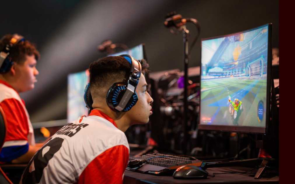 Two individuals wearing headphones playing Rocket League on monitors at a gaming event.