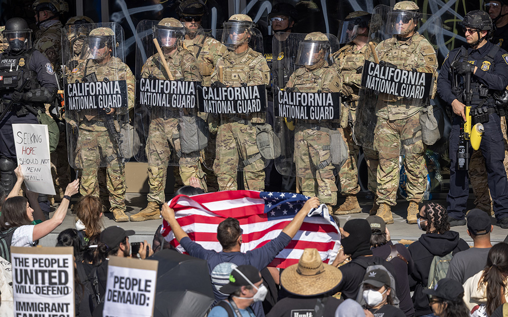 California National Guard in riot gear face protesters holding signs and an American flag.