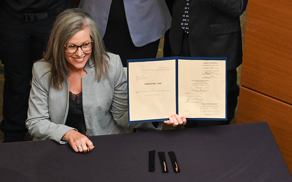 The image Gov. Katie Hobbs with shoulder-length gray hair and glasses, smiling while seated at a table. They are holding a blue-bordered document folder open, displaying two pages of text. The document is titled "SENATE BILL 1583."