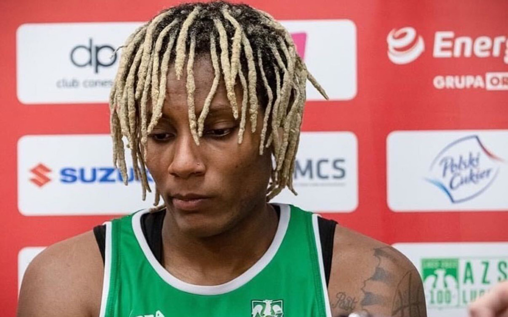 Natasha Mack with blonde dreadlocks in a green sports jersey against a red backdrop with sports logos.