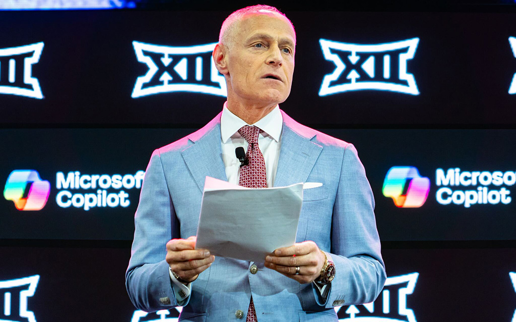 The image shows Brett Yormark standing on a stage holding a piece of paper, speaking into a microphone with a background featuring repeated logos of "XII".