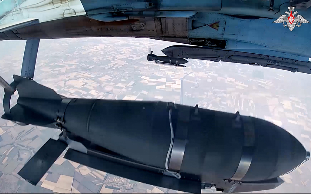 View of a large black bomb beneath an aircraft flying over patchwork fields.