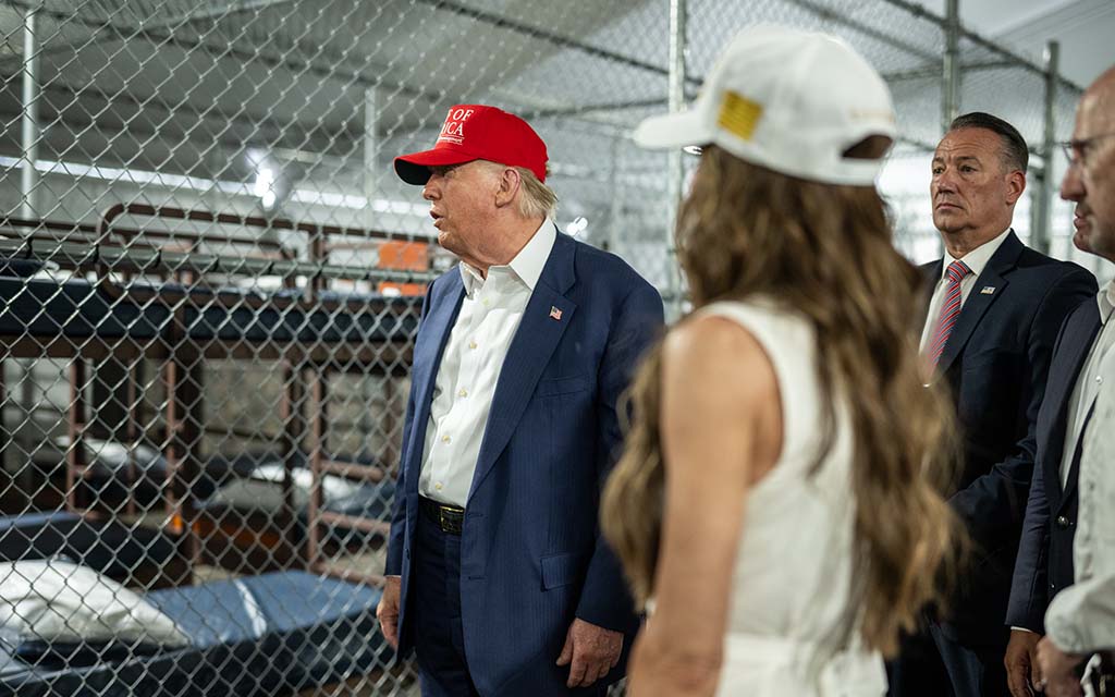 President Donald Trump and a group of people stand near a chain-link fence with bunk beds visible behind it in an indoor facility.