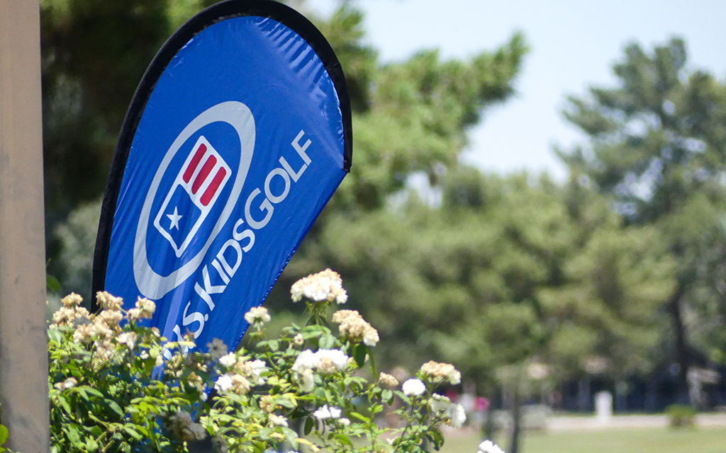 A blue teardrop-shaped banner that says U.S. Kids Golf, surrounded by white flowers and trees.